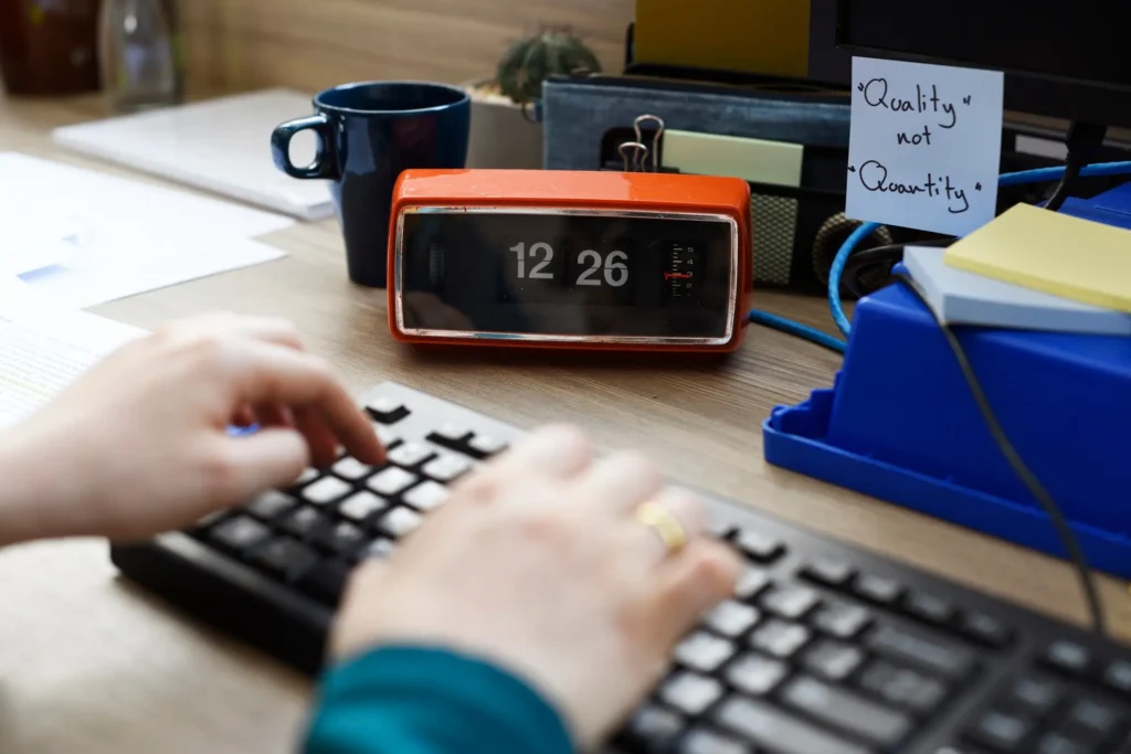 Primer plano de un espacio de trabajo con una persona escribiendo en un teclado, un reloj digital naranja que indica las 12:26 y una nota en la que se lee "Calidad, no cantidad".